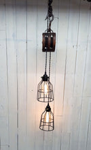 Load image into Gallery viewer, Two offset 5 1/2-inch diameter cage lamp shades housing Edison bulbs, suspended from a double wheel barn pulley, this pendant light hangs 36 inches, and has a 24-inch hanging chain. Lights are Illuminated.
