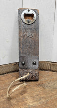 Load image into Gallery viewer, A barrel stave bottle opener with  indent cap remover and a barrel ring accent, sits on top of a whiskey barrel.  The opener has a jute rope cord at the bottom .
