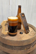 Load image into Gallery viewer, A barrel stave bottle opener with indent cap remover and a barrel ring accent, leans on an empty beer bottle, next to a mug of just poured ale, all set on top of a whiskey barrel.
