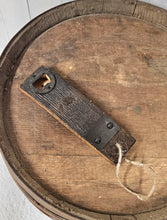 Load image into Gallery viewer, A barrel stave bottle opener with indent cap remover and a barrel ring accent, lays on top of a whiskey barrel. The opener has a jute rope cord at the bottom .
