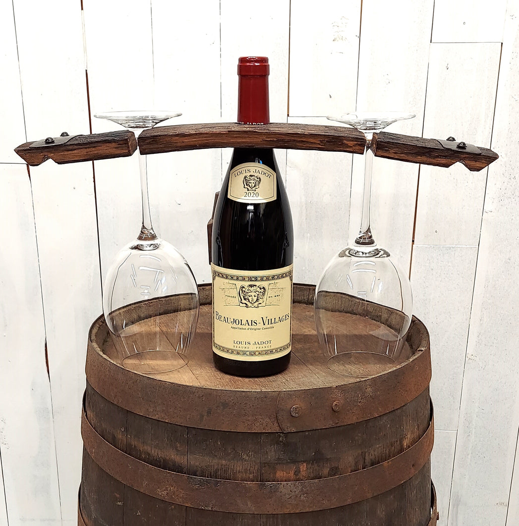 Wine bottle centered on a wooden barrel top, with a rustic barrel stave set over the bottle neck, and a stemmed wine glass hanging  on each side of the bottle.